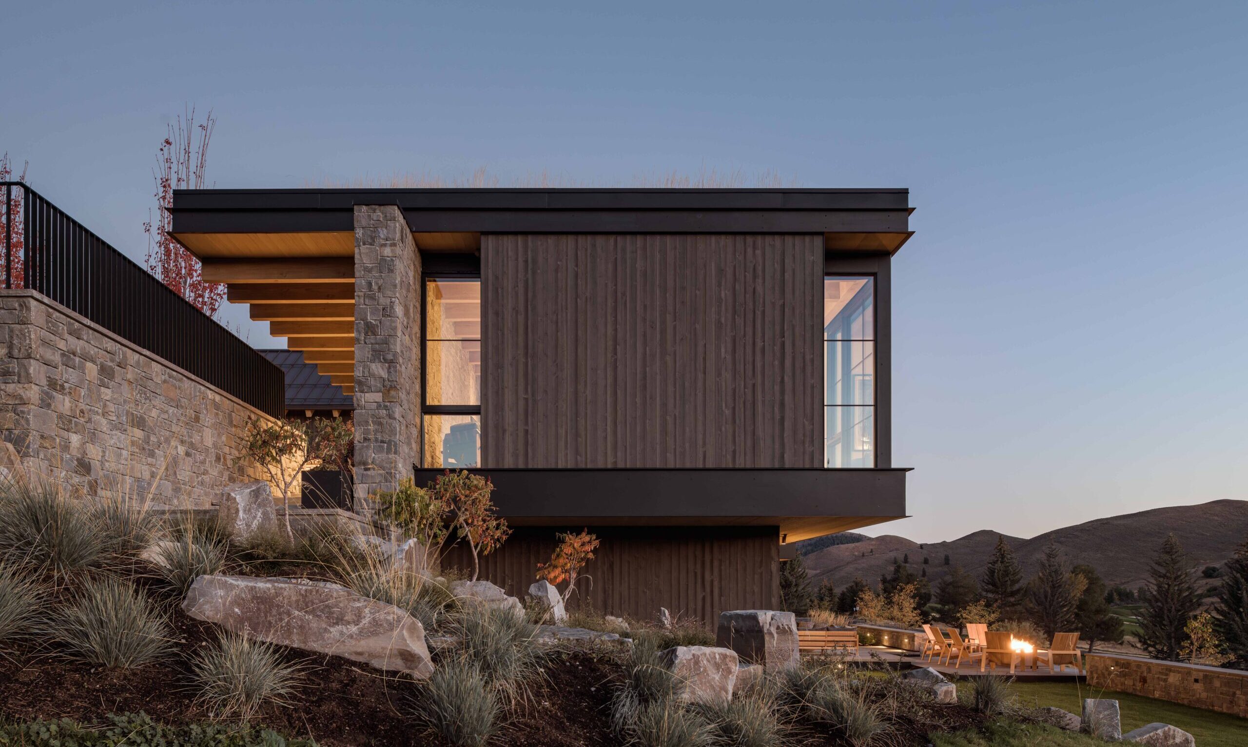 contemporary mountain home exterior, featuring dark hewn wood siding, a stone wall, and blackened steel, landscape and a patio can be seen below